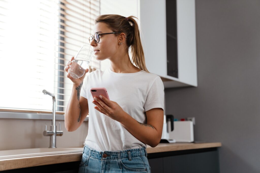 Beautiful thinking girl drinking water while using mobile phone